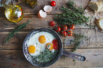 Fried eggs in a frying pan with herbs, cherry tomatoes, bread, olive oil and spices on rustic wooden background. Healthy breakfast, top view