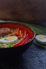 Ceramic bowl of traditional asian ramen soup with noodles, spring onion, chicken, sliced egg on dark stone table. Top view, selective focus.
