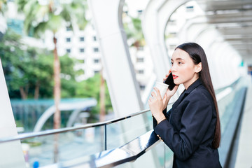 Young Asian woman with smartphone standing against street blurred building background. Fashion business photo of beautiful girl in black casual suite with phone and cup of coffee