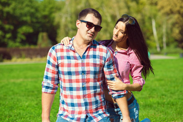 Obraz premium Young beautiful loving couple in checked shirts, jeans and sunglasses sitting on the green lawn. A girlfriend looking at her boyfriend with her hand on his shoulder. Outdoor shot