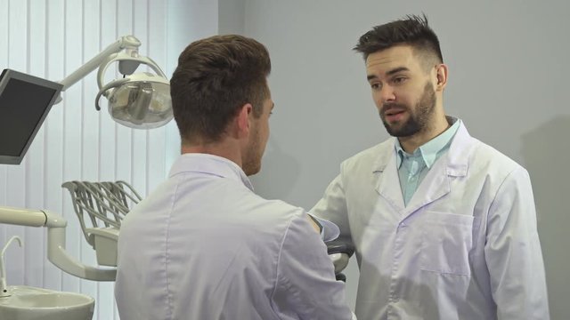 Two attractive young dentists shaking hands at the office. Handsome bearded doctor leaning on some equipment. Blond male dental specialist and his colleague discussing some working moments