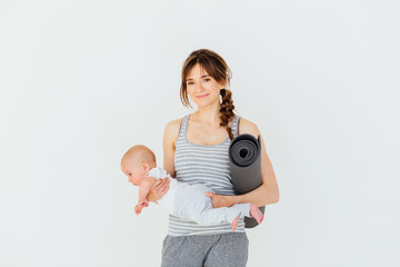 Sport, motherhood and active lifestyle concept - happy charming sporty mother holding cute little baby boy in one hand and yoga mat in the other while standing on white background.