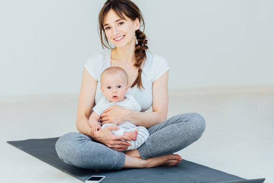 Sport, Motherhood And Active Lifestyle Concept - Happy Mother And Little Baby Boy Sitting On Yoga Mat And Get Ready To Start Workout At Home Over Light Background.