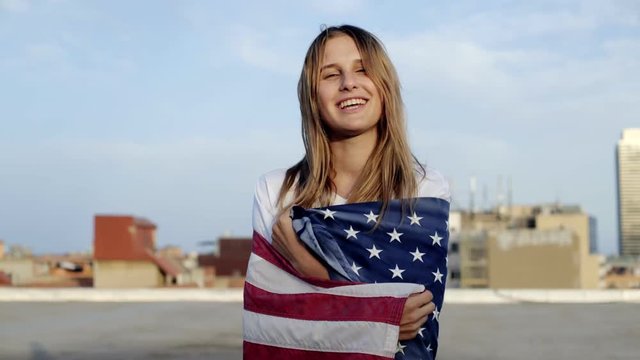 Attractive sexy blonde young woman wrapped in american flag smiles into the camera, happy and laughing, excited for summer adventures and holidays, concept freedom, youth, millennial, hipster