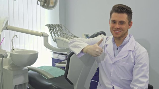 Handsome caucasian dentist posing with layout of human teeth at the office. Attractive blond doctor raising his thumb up. Young male dental specialist gesturing good health
