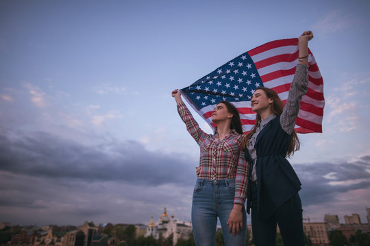 American teens with flag travel around the world. Happy girls on roof. Teenager sisters on vacation celebrate the independence day on sky background