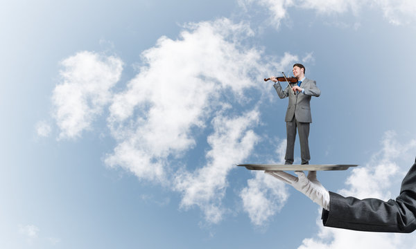 Businessman on metal tray playing violin against blue sky background