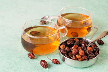  Rose hips, dried fruit in a metal bowl and freshly prepared tea in clear glass mugs on a light green background.
