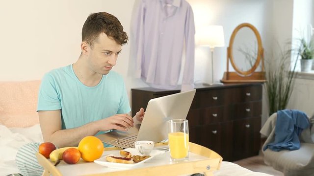 Young man smiling to the camera while using laptop and having breakfast in bed, steadycam shot

