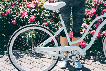 Female legs in blue sneakers and a stylish bicycle against a background of bushes with roses