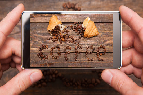 Mobile conceptual photography. Phone in hands taking photo of insctiption made by coffee beans and sweet dessert on wooden background for social network, top view