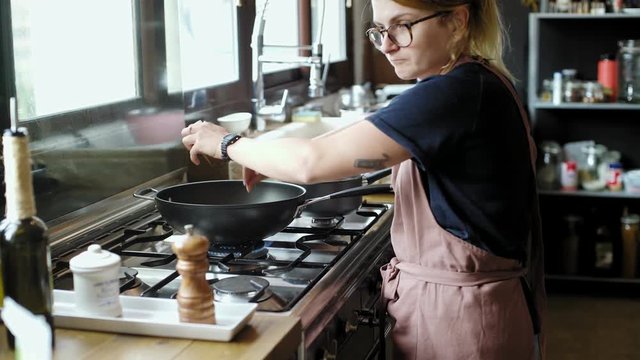 Young petite woman prepares dinner or lunch in her home industrial chic kitchen on stainless steel stove, wears apron and adds exotic herbs and spices to fresh ingridients on stir fry wok pan