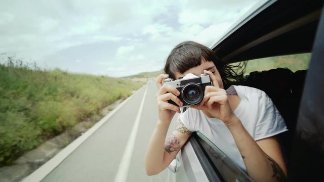 Pretty authentic millennial woman hangs out of backseat window, makes photos on her vintage film camera, smiling and her brunette hair blowing in wind, concept speed, getaway, vacation, summer