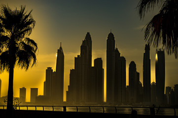 Artistic, black and yellow panorama of skyscrapers in Dubai from promenade with palm trees at the foreground at sunrise. UAE