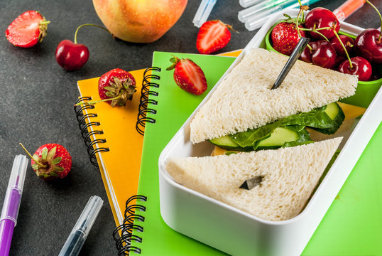 Back to school. A hearty healthy school lunch in a box: sandwiches with vegetables and cheese, berries and fruits (apples) with notebooks, colored pens on a black table.