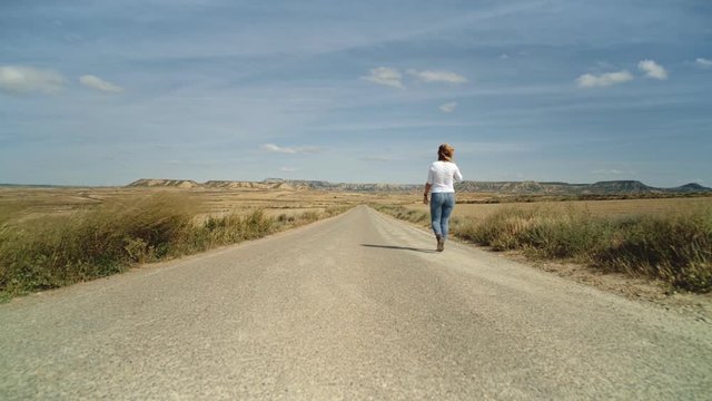 Lonely urban traveller nomad blonde woman in raw denim jeans and simple white shirt walks away into distance in empty and wild desert environment, looks for freedom and independence