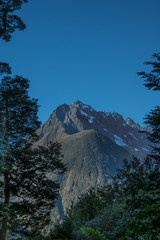 Fiordland National Park, New Zealand - March 16, 2017: Tall, dark, snow-covered mountain towers over The Chasm area under dark blue sky. Some green trees as frame.