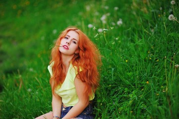 Naklejka na ściany i meble Portrait of young attractive girl with bright red hair sitting on green grass with a weary look, closeup.
