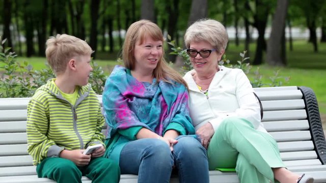 Happy grandmother, mother and son sitting on the bench and talking in summer park