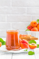 Apricot jam in a jar and fresh fruits with leaves on white wooden table, breakfast