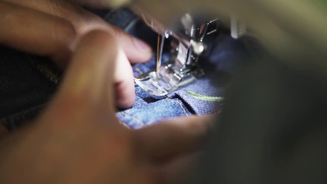 Unrecognizable female tailor hands use sewing machine to stitch patch with rose image to denim jeans pocket in work shop close up slow motion