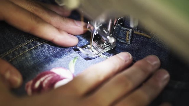 Unrecognizable woman tailor hands use sewing machine to stitch patch with rose image to denim jeans pocket in work shop close up slow motion