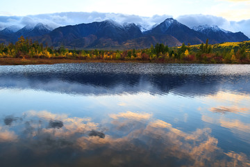 Pink cloud reflections in smooth water surface, colorful forest and mountain peaks capped with snow, autumn landscape on sunset