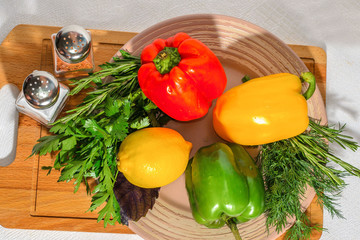 Fresh bright vegetables: Bulgarian pepper, herbs, lemon, on a ceramic plate, on a wooden cutting board