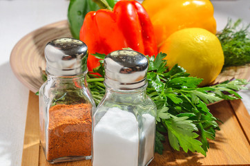 Salt shaker and pepper with fresh bright vegetables: Bulgarian pepper, herbs, lemon, on a ceramic plate, on a wooden cutting board