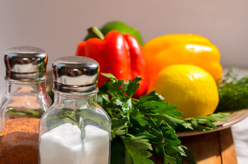 Salt shaker and pepper with fresh bright vegetables: Bulgarian pepper, herbs, lemon, on a ceramic plate, on a wooden cutting board