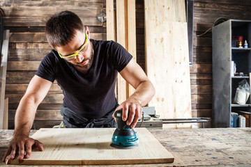 Young man builder carpenter equals polishes wooden board with a  random orbit sander  in the...
