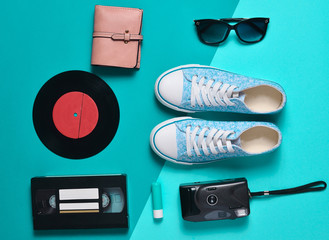 Keds, vinyl records, video cassettes, plastic vintage camera, purse are laid out on a blue neon ...