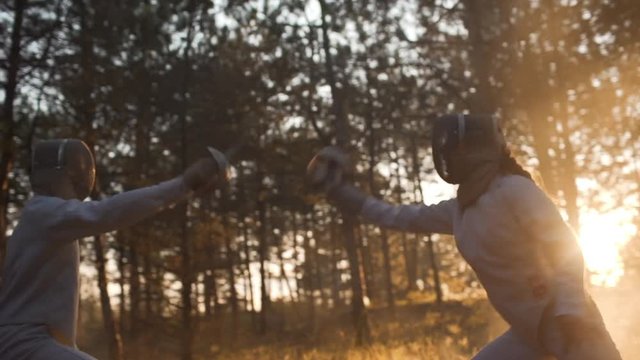 Two sportsmen fencing in the forest wearing full outfit in slowmotion. Footage with haze and sun flare