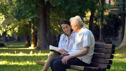A nanny, a nurse, caring for the elderly a girl (woman) and grandfather sit on a paperback and read a book, in the park. Concept boarding house, sanatorium, house for the elderly, help for the elderly