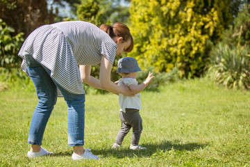 Fourteen months old baby girl making her first steps; walking on her own on the lawn on a sunny day; mother helping her by holding her from behind - Powered by Adobe