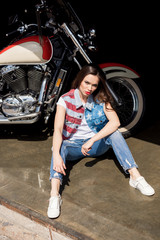 Fototapeta na wymiar Stylish young brunette woman in denim vest sitting near motorcycle and looking at camera