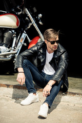Fototapeta na wymiar Handsome stylish young man in leather jacket and sunglasses sitting on concrete curb near motorbike