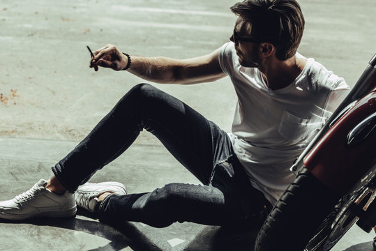 Handsome young man in sunglasses holding cigarette while sitting near motorcycle and looking away