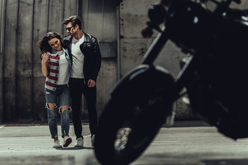 Stylish young couple walking embracing near motorbike standing outdoors