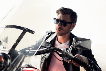 Obraz na płótnie Canvas Handsome stylish young man in sunglasses and leather jacket sitting on motorcycle and looking away