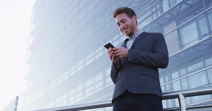 Man sms texting using app on smart phone in city business district. Young business man using smartphone smiling happy wearing suit jacket outdoors. Urban male professional in her 20s. RED EPIC.