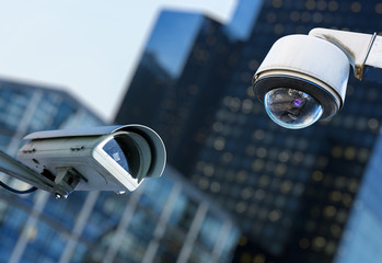 two cctv security camera in a city with blury business building on background - obrazy, fototapety, plakaty