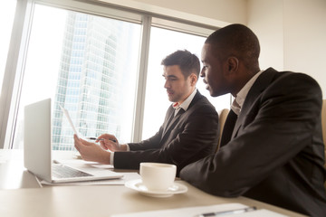 Fototapeta premium Side view of two multiracial business partners in formal wear discussing document on meeting sitting at desk, black and white businessmen studying contract terms, explaining idea, planning project