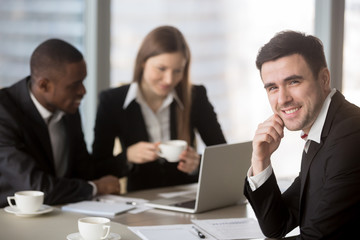 Cheerful businessman wearing suit looking at camera sitting at desk with international partners, experienced team leader holding business meeting for subordinates at background, consulting services