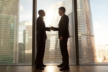 Two smiling young black and white businessmen handshaking standing near big window with city buildings outside, confident caucasian and afro american partners form good relations, reach agreement