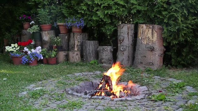 A stone fireplace with fire in a green traditional garden. Large wooden blocks with flowers in the background. 