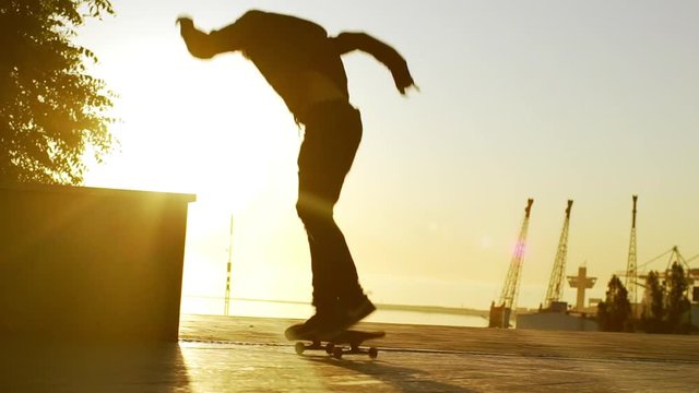 Caucasian male skateboarder performing ollie trick from parapet with sea port on background. Sunlit footage in slowmotion with sun flare