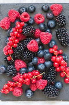 Mix of berries raspberries red currants blueberries and blackberries on black slate board. White stone background.  Top view.