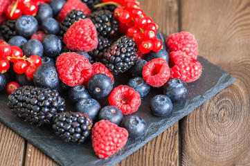 Mix of berries raspberries red currants blueberries and blackberries on black slate board. Wooden background.  Copy space.
