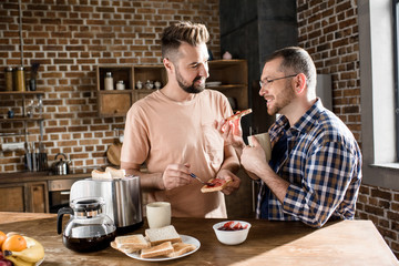 Happy gay couple eating toasts with jam in kitchen at morning